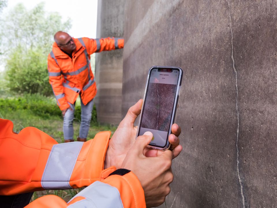 Two hands are holding a phone near a crack in the wall. You can see the Crack Width Analysis app, which is scanning the wall. In the background, another person is checking the wall.