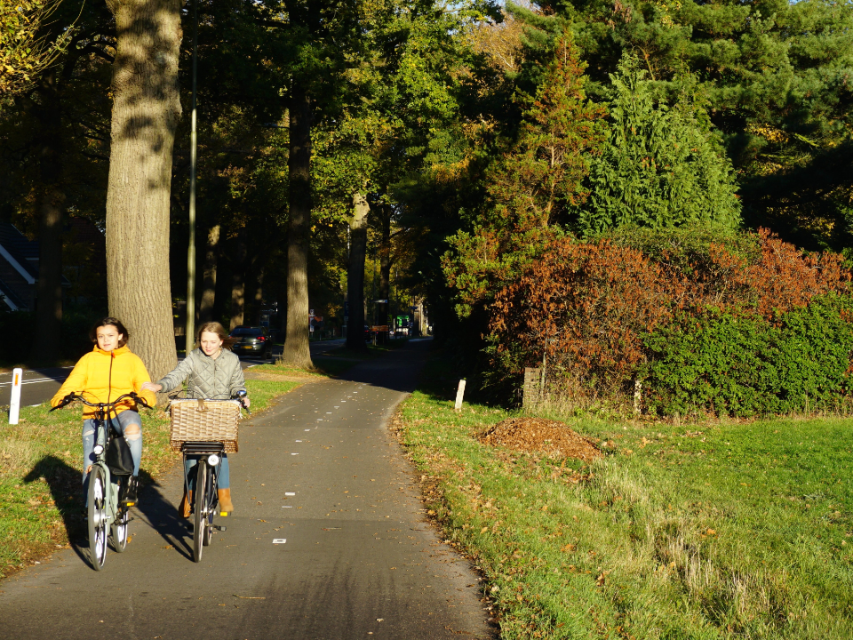 Een foto van 2 jonge kinderen op de fiets over een geasfalteerd fietspad naast een weg.