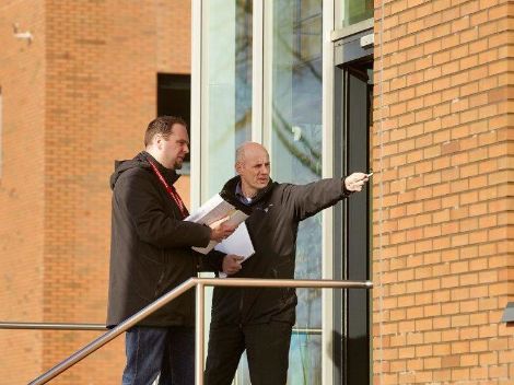 Two men are standing in front of a building. One has a memo in his hand, the other points to something outside the picture.