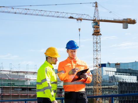 Two men wearing helmets and yellow vests are looking at a memo. They are standing on a building site. In the background is a large crane.