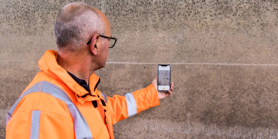 Een man houdt zijn telefoon bij een muur. Hij test de Crack Width Analysis-app.