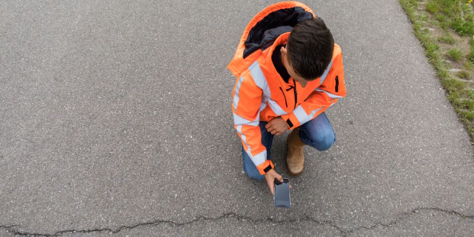 A man sits near a crack in the asphalt. He checks the crack with the Crack Width Analysis app.