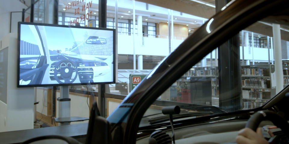 Een foto van een auto in een gebouw. Op een scherm voor de auto zie je wat de auto zou zijn als die op de weg zou rijden.
