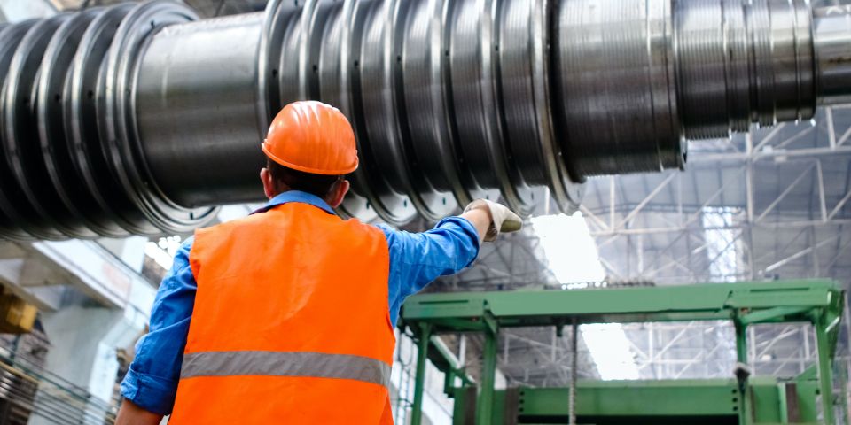 A man wearing a vest and an orange helmet stands by a large pipe in a factory.