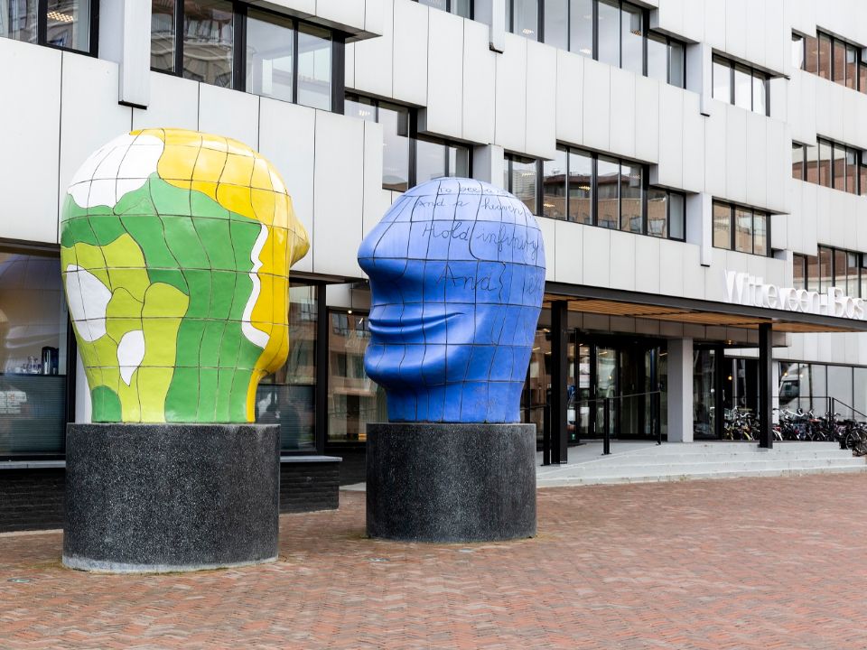 Zij aanzicht van het hoofdkantoor in Deventer. Het pand is wit met zwarte ramen en er staat een kunstwerk voor van twee grote hoofden.